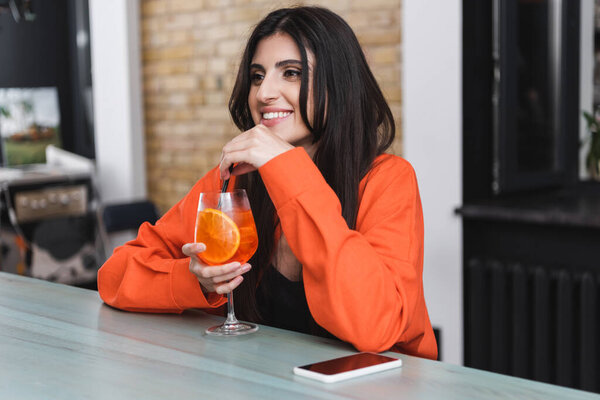Positive woman holding drinking straw in cocktail near mobile phone in cafe 