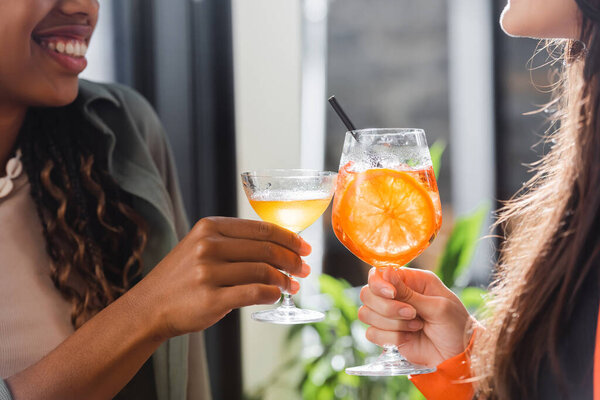 Cropped view of multiethnic girlfriends clinking cocktails in cafe 