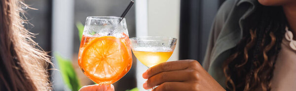 Cropped view of multiethnic girlfriends clinking cocktails in cafe, banner 