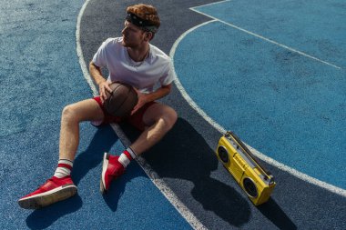 full length of man in sportswear sitting on court with ball and tape recorder