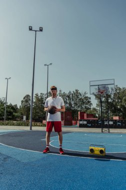 full length of sportive man with ball standing on basketball court near boombox
