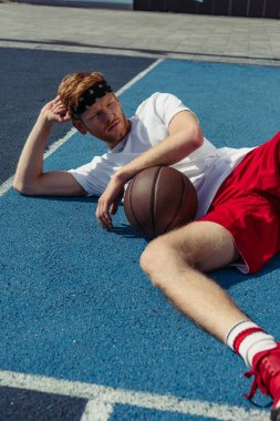 redhead basketball player in sportswear lying on court near ball and looking away