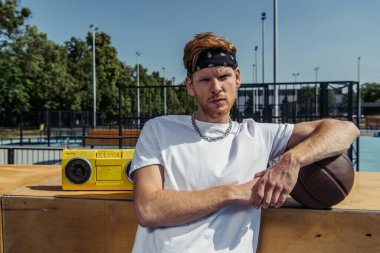 sportive man in bandana and necklace looking at camera near ball and tape recorder
