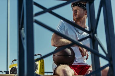young basketball player sitting with ball and boombox near blurred fence
