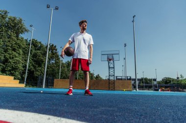 full length of basketball player in sportswear standing with ball on modern court