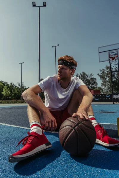 young basketball player in red sneakers sitting on court near ball