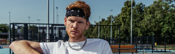 young sportive man in bandana and necklace looking at camera outdoors, banner