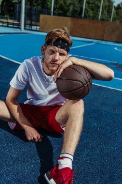 man in bandana and red shorts sitting with ball on modern court