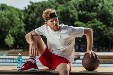 sportive man sitting near ball and sports bottle and looking away
