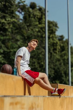 sportive man with closed eyes sitting on stadium near ball and sports bottle
