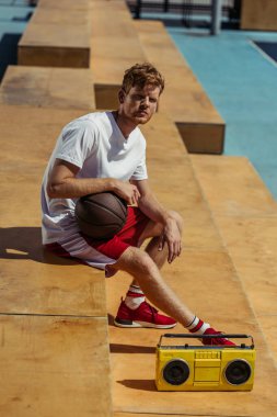 young sportsman sitting with ball near boombox on stadium