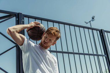low angle view of redhead man in white t-shirt standing with ball near fence 