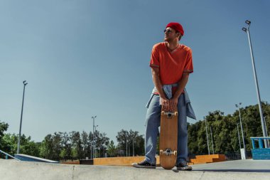 full length of man in orange t-shirt and jeans standing with skateboard in park