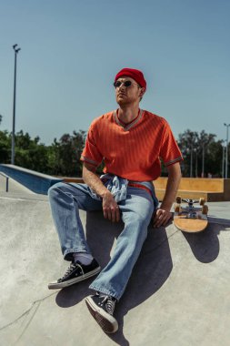 man in trendy clothes and sunglasses sitting on ramp near skateboard
