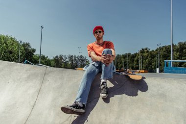 full length of stylish skateboarder in gumshoes sitting on ramp in skate park