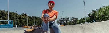 fashionable man in beanie and sunglasses sitting near skateboard in park, banner