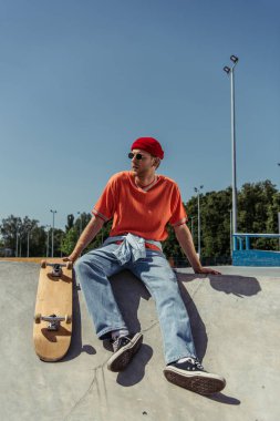 full length of fashionable man in gumshoes and sunglasses sitting on ramp near skate