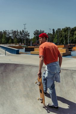 young man in orange t-shirt and jeans walking on ramp with skateboard