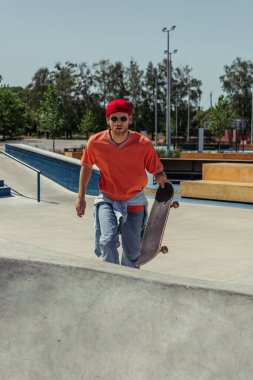young man in red beanie and sunglasses walking on ramp with skateboard