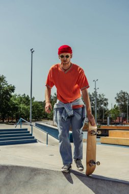 full length of man in jeans and orange t-shirt holding skateboard in park