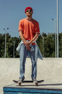 full length of young man in jeans and sunglasses standing on skate in park