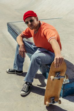 man in sunglasses and red beanie sitting with skate and looking at camera