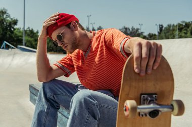 tired man touching beanie while sitting with skateboard in park