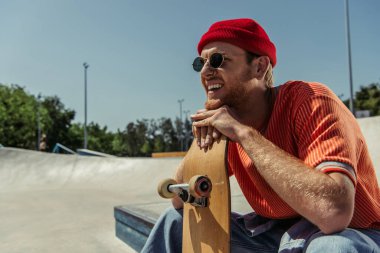trendy man in sunglasses leaning on skateboard and looking away