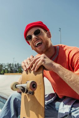 excited man in sunglasses laughing near skate outdoors