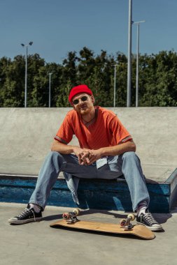 trendy man in sunglasses and red beanie sitting near skate and smiling at camera
