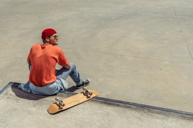 back view of man in orange t-shirt and red beanie sitting near skateboard on ramp