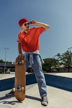 full length of man adjusting trendy sunglasses while standing with skate in park