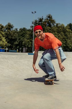 excited man in red beanie and sunglasses riding skateboard in skate park