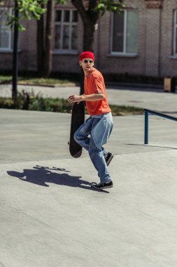 stylish man in sunglasses looking back while running on street with skateboard
