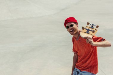 excited man in red beanie and orange t-shirt holding skateboard and looking at camera