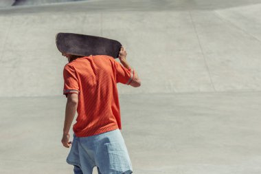 back view of man in orange t-shirt carrying skateboard on shoulders 
