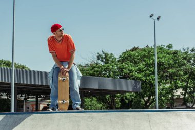 full length of man in trendy outfit standing with skateboard and looking away