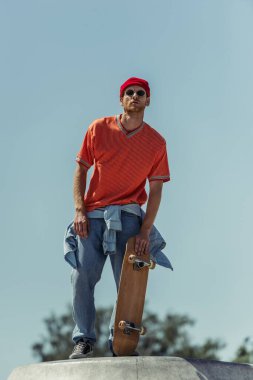 low angle view of man in sunglasses and orange t-shirt holding skateboard outdoors