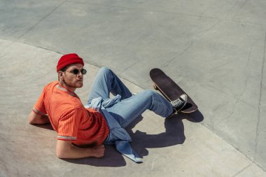 high angle view of man in sunglasses and trendy clothes sitting on ramp near skateboard