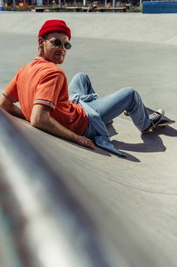fashionable skateboarder looking at camera while sitting on ramp on blurred foreground
