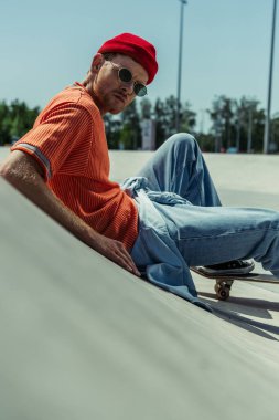 trendy skater in sunglasses sitting on ramp and looking at camera