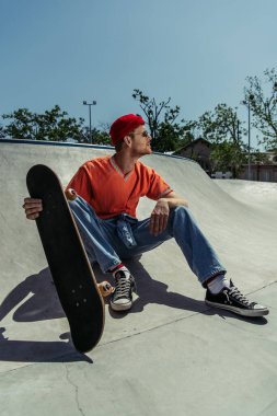 young man in trendy clothes sitting on ramp with skateboard