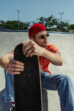 young and trendy man in sunglasses sitting with skateboard and looking away