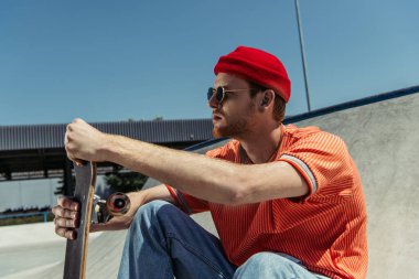 young man in red beanie and sunglasses sitting with skate outdoors