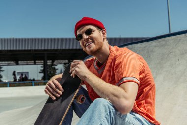 happy man in stylish outfit sitting with skateboard outdoors