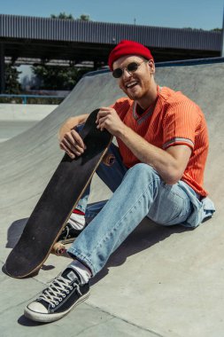 happy and fashionable man sitting with skateboard and smiling at camera