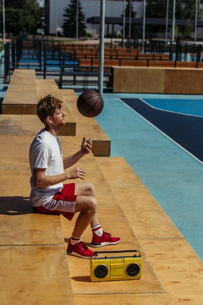 side view of sportive man playing with ball while sitting on stadium near record player