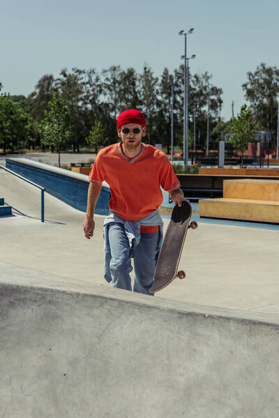 Молодой человек в красной шапочке и солнцезащитных очках ходит по пандусу со скейтбордом