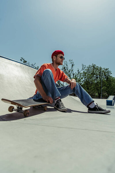 full length of trendy man in gumshoes and sunglasses sitting near skateboard