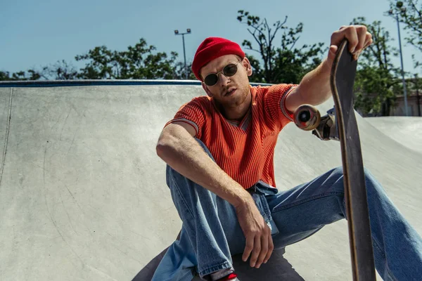 stylish man in sunglasses sitting with skateboard and looking at camera